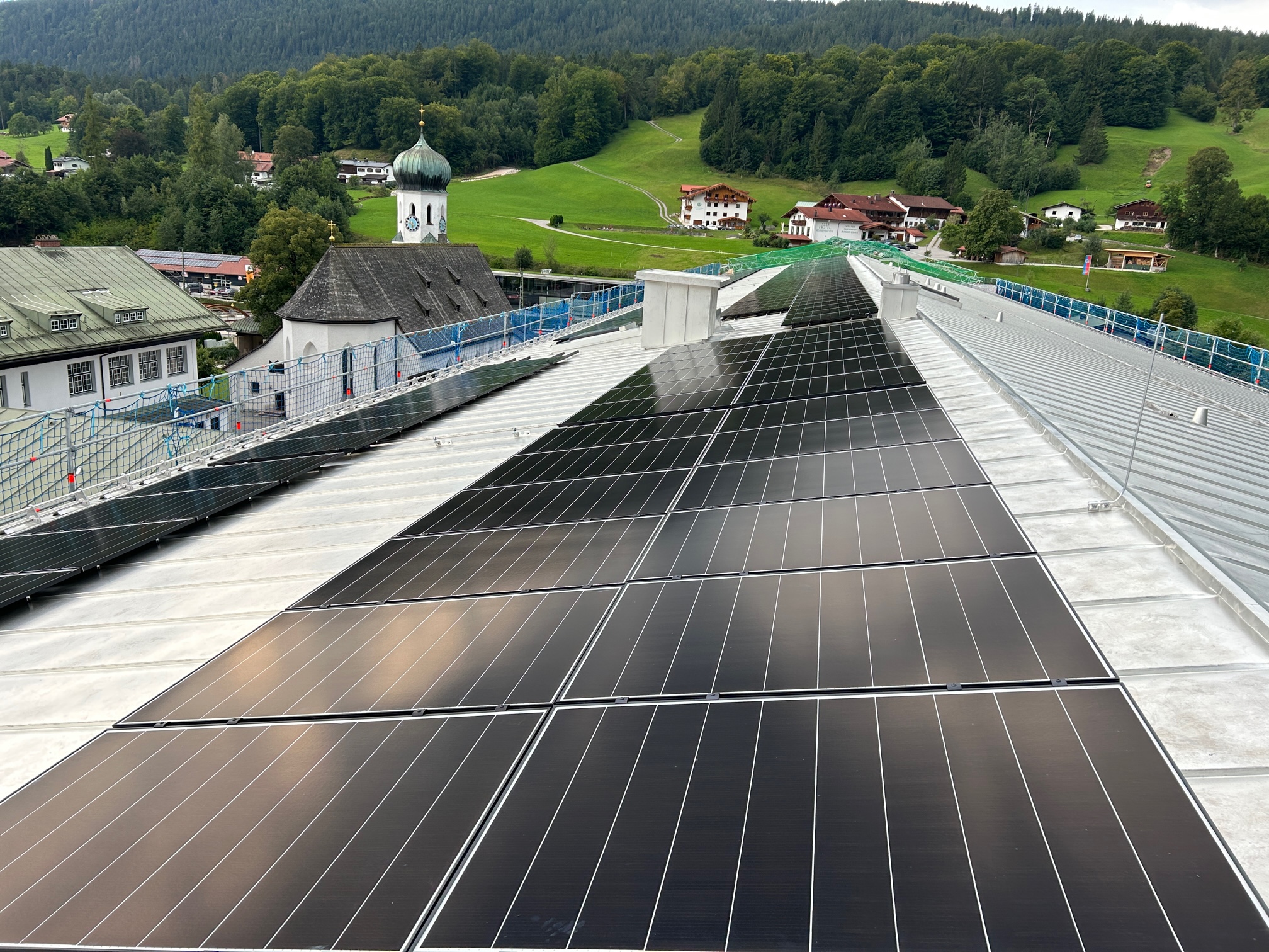 Die PV-Anlage auf dem Dach der Mittelschule Bischofswiesen mit Kirchturm und Hügeln im Hintergrund.